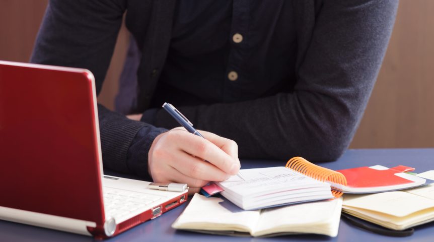 Redator anotando palavras-chave em seu caderno e criando um conteúdo para blog em seu notebook.